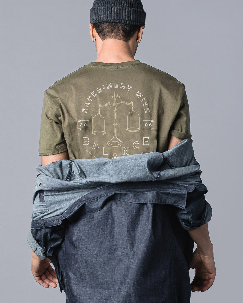 back of man wearing olive tee with "experiment with balance" balancing scale design 