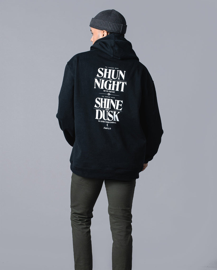 back of man wearing black hoodie with "to those who shun the night we tip our hat. To those who shine after dusk we offer a warm embrace"